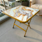 Mesa plegable multiusos cuadrada de 80x80 cm con acabado tipo mármol y estructura dorada, vista en perspectiva superior.