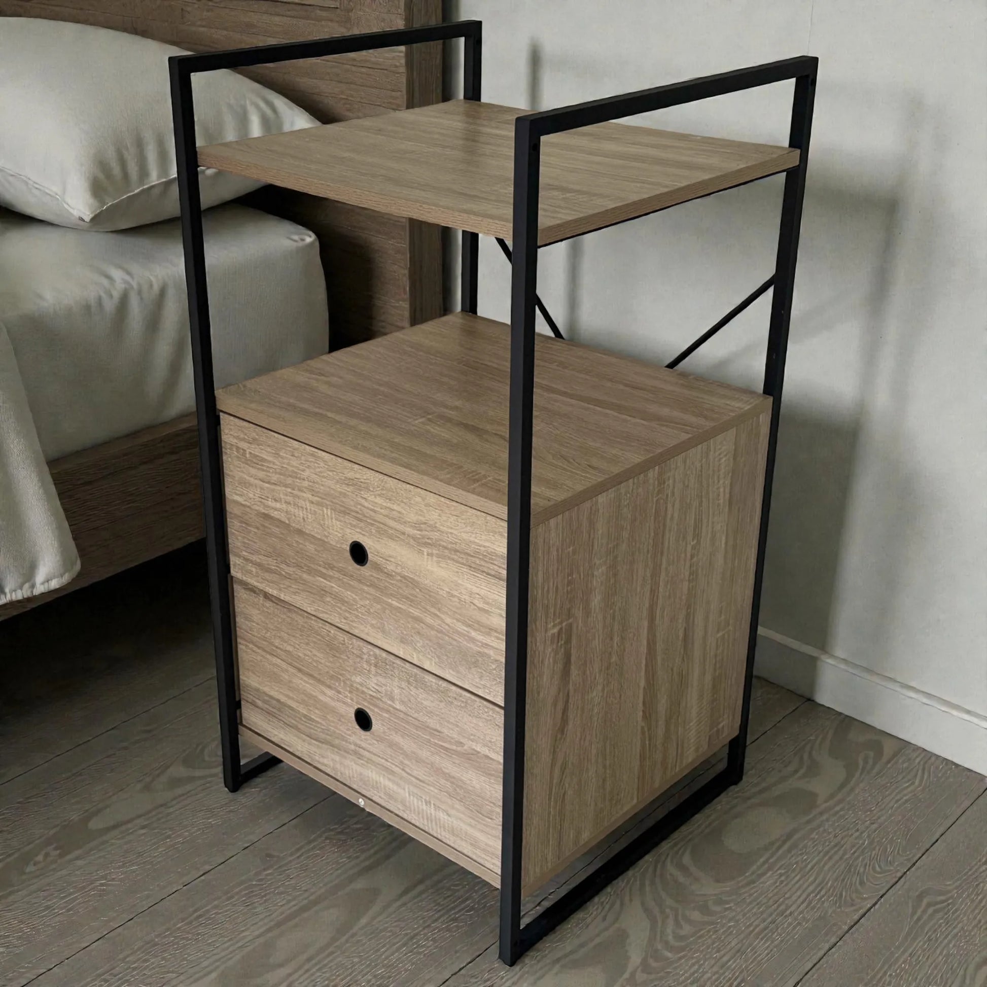 Mesita de noche Flexi Storage Studio de 60 x 58 x 113 cm colocada junto a la cama, con estructura metálica negra, paneles color roble, repisa superior y dos gavetas para almacenamiento en dormitorio.