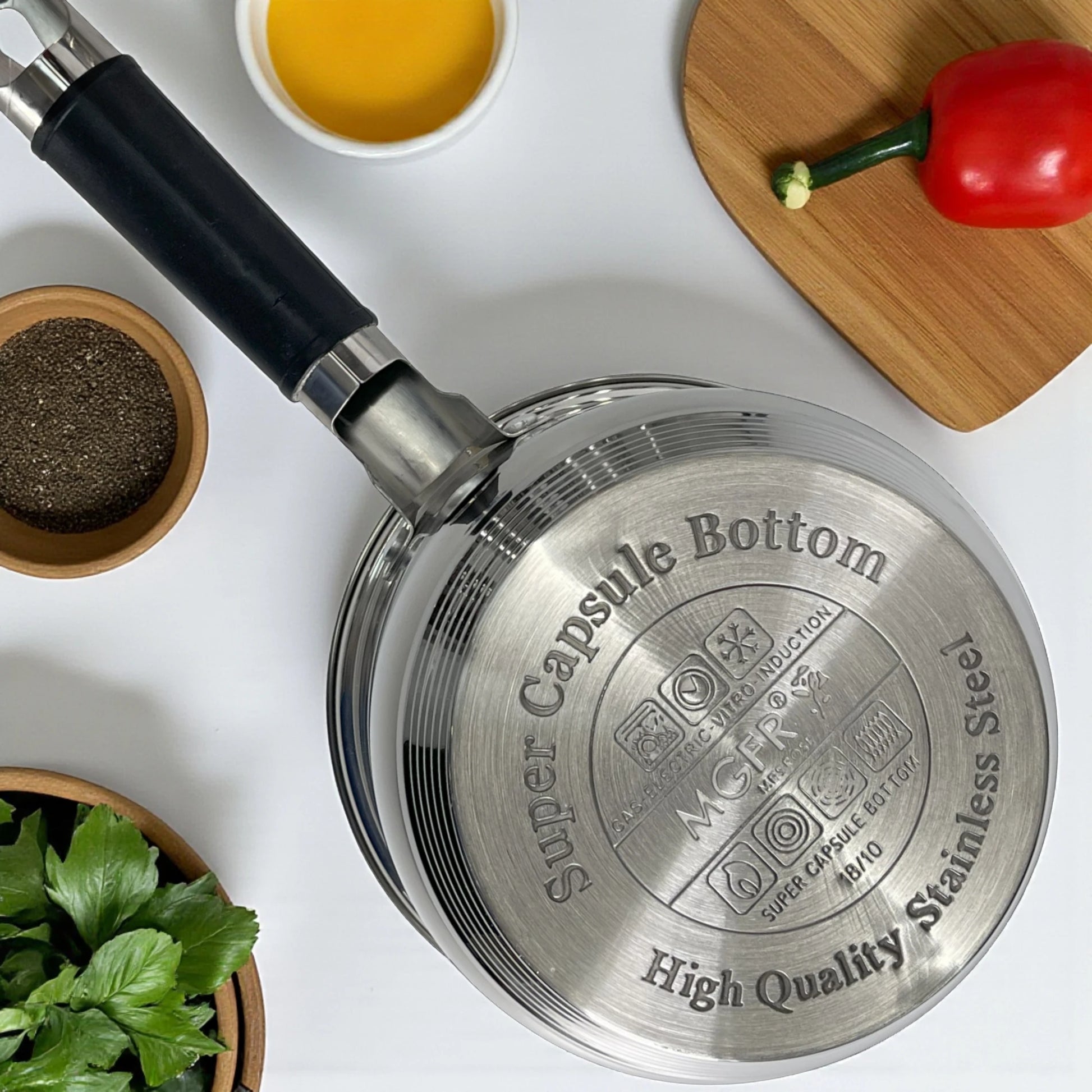 Vista superior de la sartén MGFR MR-290G de 24 cm con base “Super Capsule Bottom” de 7 capas en acero inoxidable 18/10; mango negro a la izquierda, encimera blanca con bowls de pimienta, salsa y pimiento rojo.