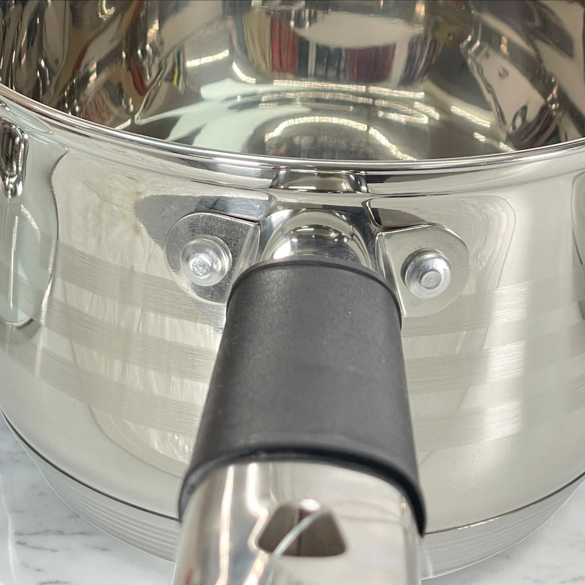 Primer plano del cazo MGFR 16 x 10.5 cm en acero inoxidable acabado espejo; unión del mango negro con doble remache y borde pulido sobre encimera de mármol.