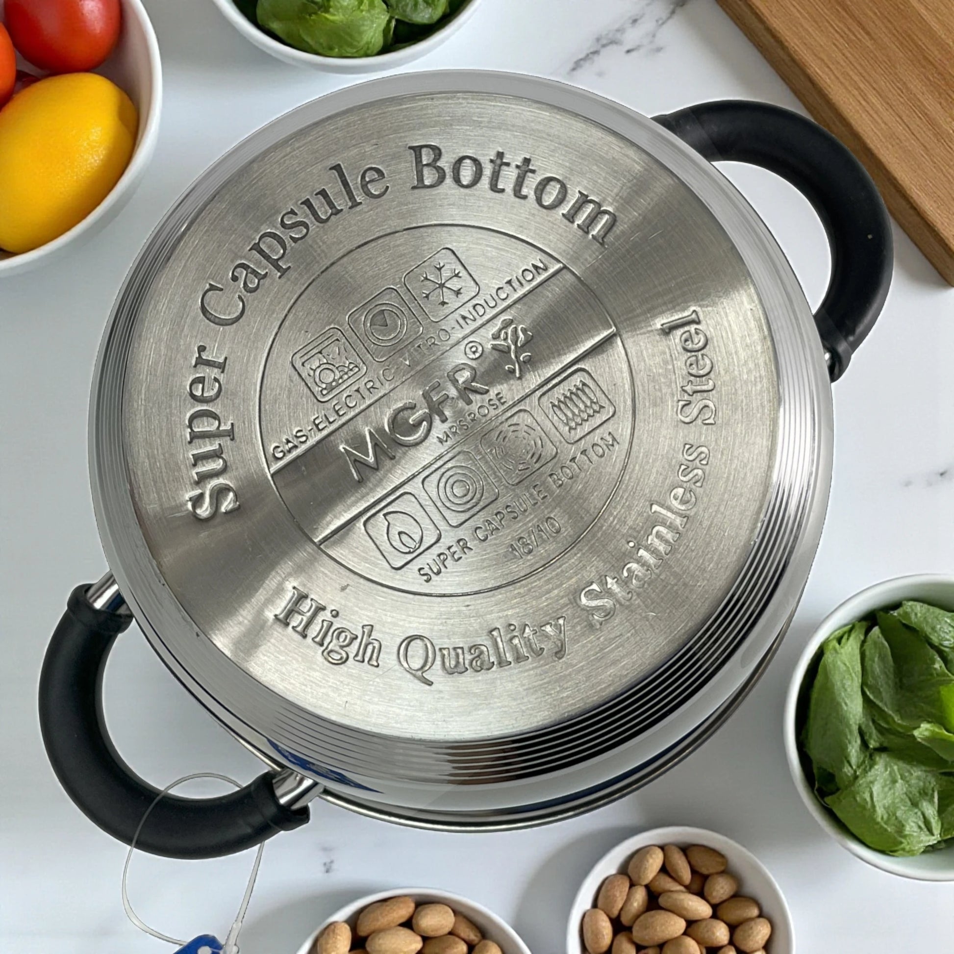 Vista superior de la base de olla MGFR 24 cm con grabado “Super Capsule Bottom” de 7 capas; acero inoxidable 18/10, compatible con gas, eléctrica, vitro e inducción; asas negras visibles y bowls con limones, almendras y hojas verdes alrededor.