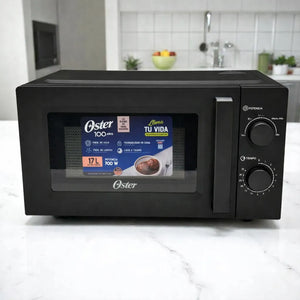 Microondas Oster OGW1702 color negro, 17 litros y 700 W, vista frontal con puerta cerrada, ventana con etiqueta del producto, perillas mecánicas negras de potencia y tiempo con marcaciones en blanco, sobre encimera blanca en cocina.