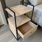 Mueble auxiliar Flexi Storage Studio con estructura metálica negra y paneles roble, visto en ángulo superior, mostrando uno de los cajones inferiores abiertos y la repisa superior, ideal para guardar ropa o accesorios.
