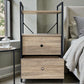 Mueble auxiliar Flexi Storage Studio con dos gavetas y una repisa, color roble con estructura negra, ubicado junto a la cama; el cajón inferior está abierto mostrando espacio para ropa, zapatos o textiles.