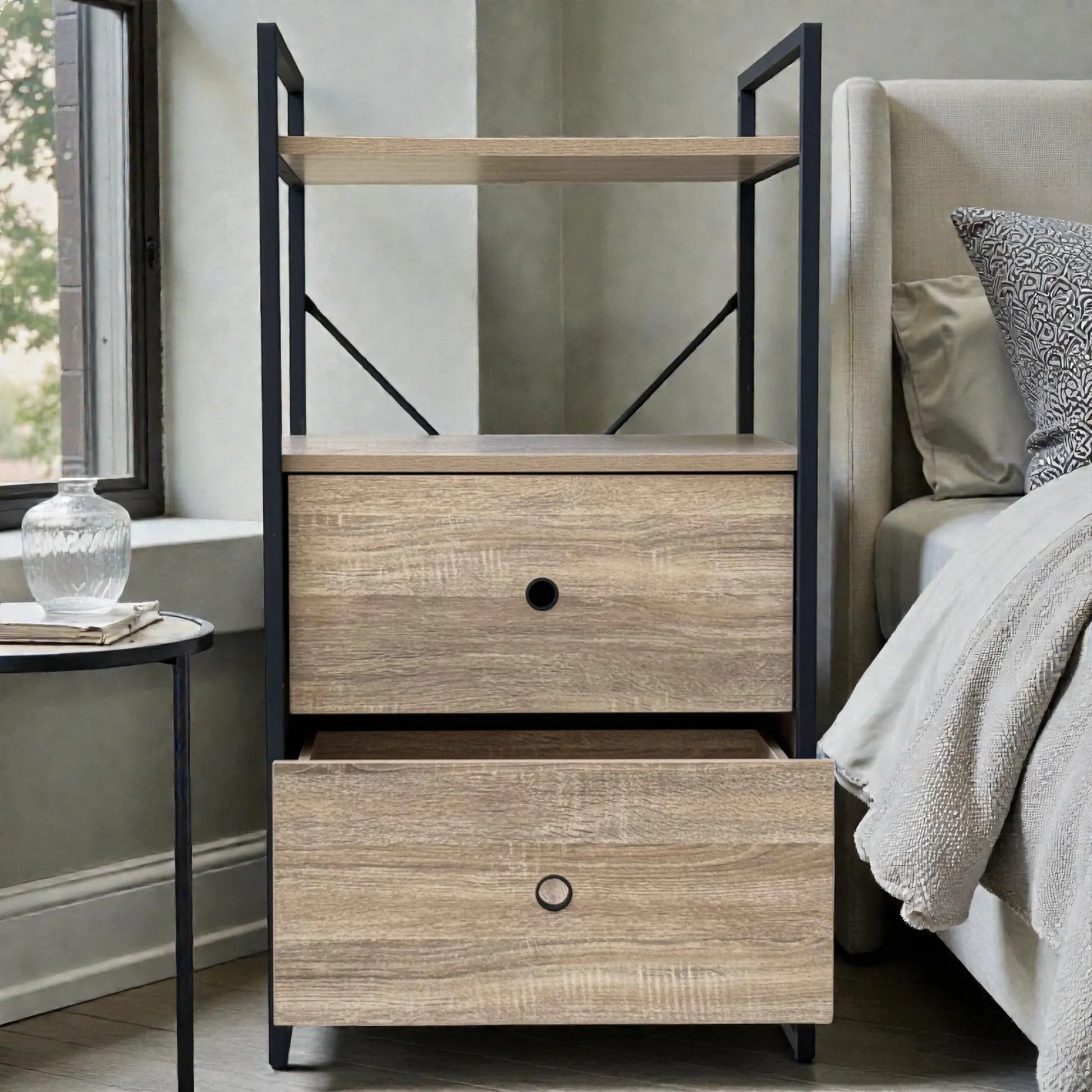 Mueble auxiliar Flexi Storage Studio con dos gavetas y una repisa, color roble con estructura negra, ubicado junto a la cama; el cajón inferior está abierto mostrando espacio para ropa, zapatos o textiles.