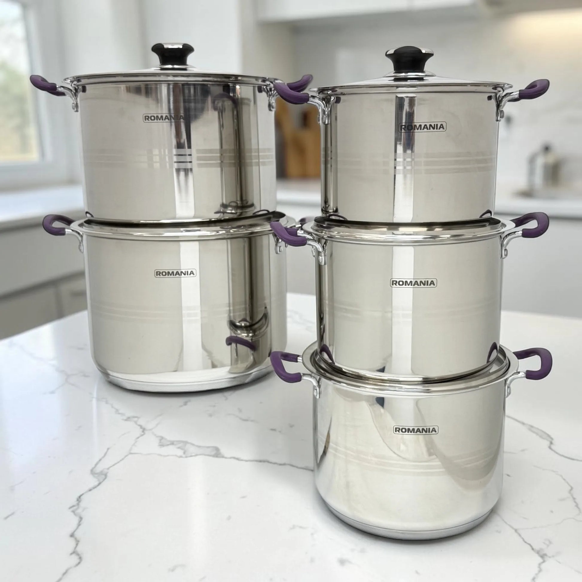 Vista frontal de varias ollas Romania en acero inoxidable con mangos púrpura, apiladas sobre encimera blanca.