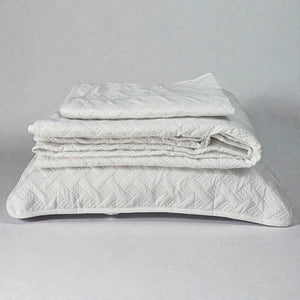 Foto tipo catálogo/estudio sobre fondo neutro gris. El set está doblado y apilado, mostrando grosor y costuras. Se aprecia el acolchado y el borde (ribete) blanco.