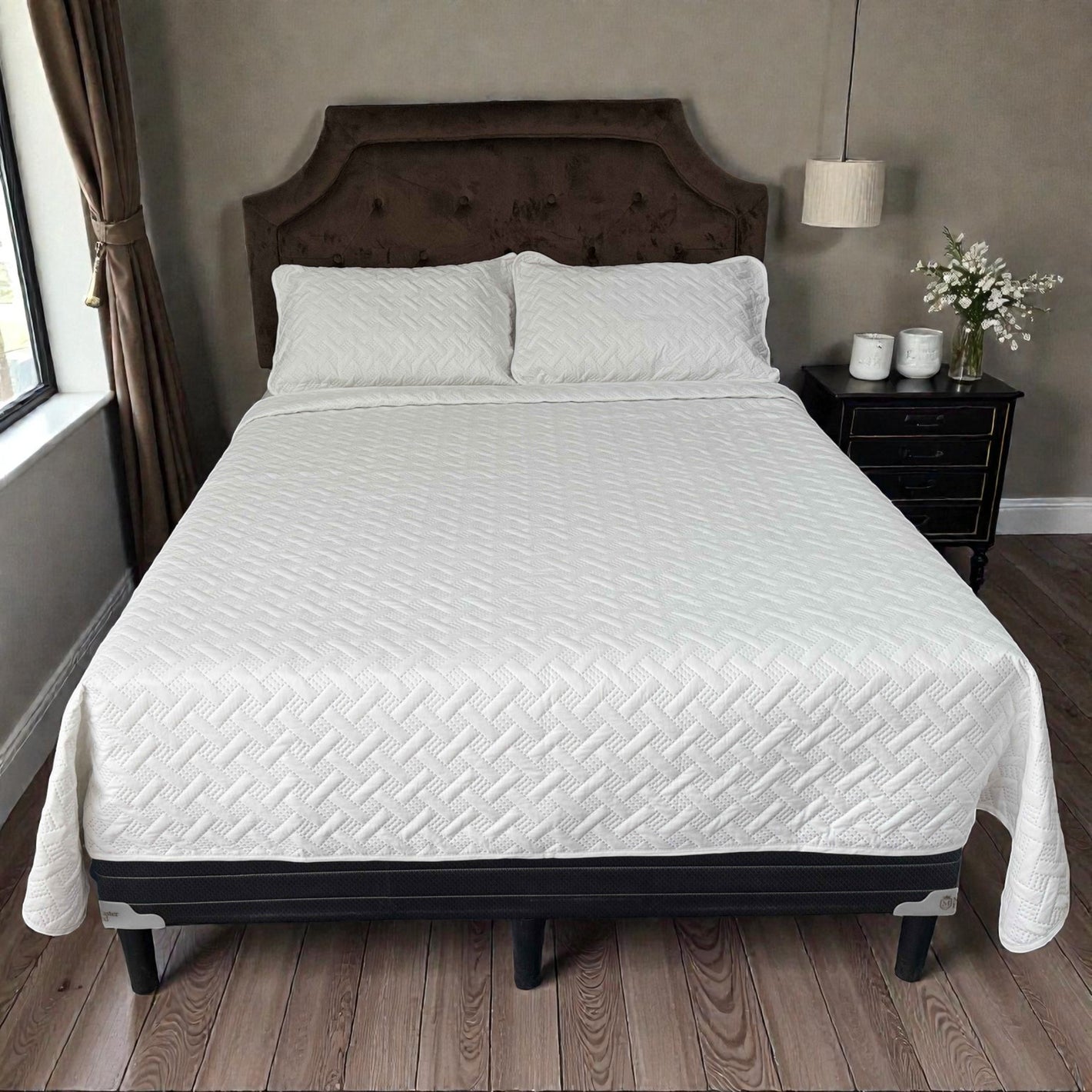 Toma frontal centrada de la cama completa. Se ve la colcha blanca acolchada con patrón geométrico tipo “trenzado/espiga” y 2 fundas a juego. Fondo tipo habitación: cabecera café, mesa de noche a la derecha, lámpara colgante y ventana a la izquierda.