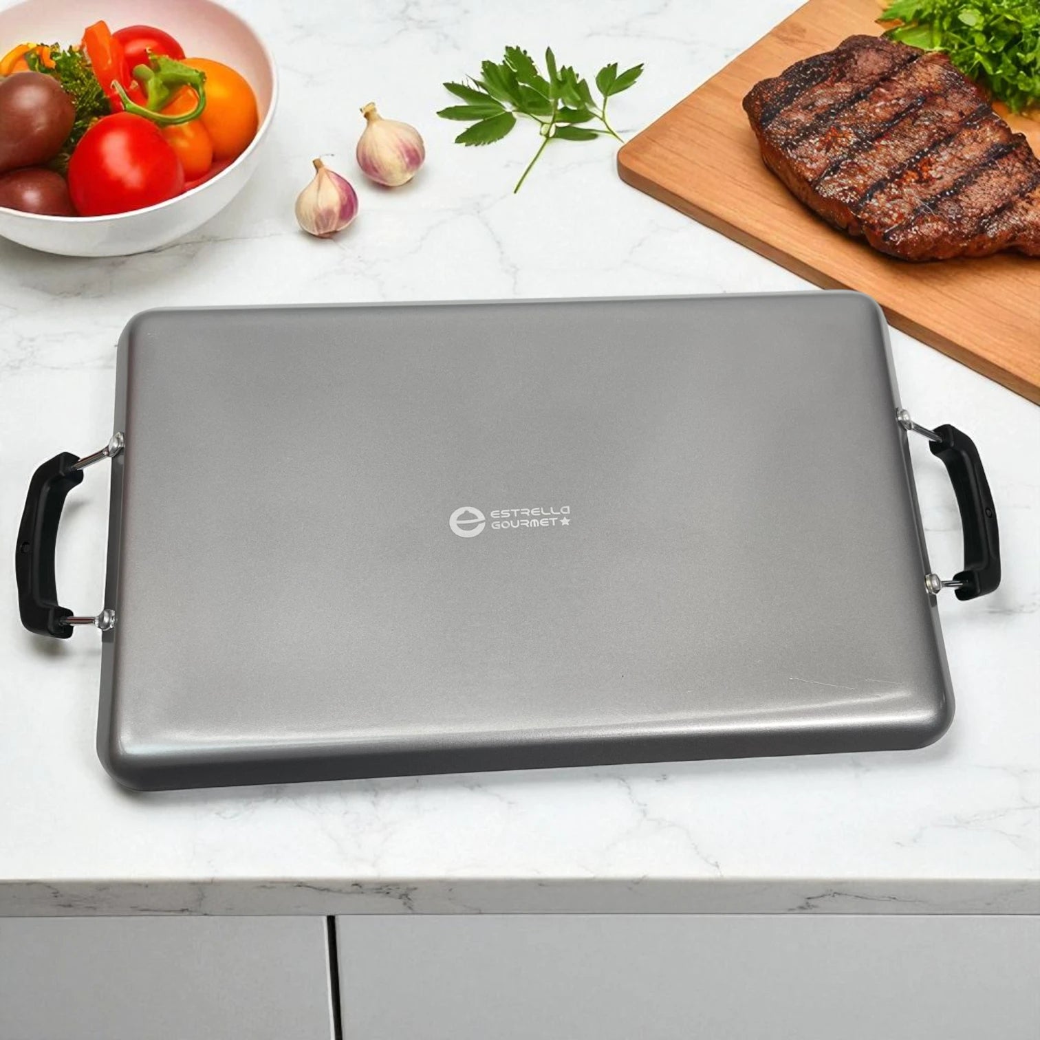 Plancha Estrella Gourmet de doble quemador vista desde el reverso, colocada sobre mesa con vegetales frescos y carne asada.