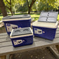 Set de hieleras Princeware New Icy Chiller: hielera 14L con tapa levantada mostrando interior, junto a hielera 30L y 6L en color azul con detalles blanco/gris sobre mesa de picnic en parque.