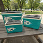 Set de 3 hieleras térmicas Princeware New Icy Chiller turquesa con detalles blanco/gris (6L, 14L y 30L) sobre mesa de picnic en parque, con porta vasos en la tapa.