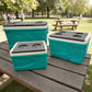 Tres hieleras Princeware New Icy Chiller del set 3409-C1PB: 6L al frente, 14L a la derecha y 30L atrás, color turquesa con detalles blanco/gris, en mesa de picnic con parque de fondo.