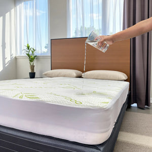 Mano vertiendo agua sobre el protector de colchón impermeable Scarlett Classics Bamboo Sensation, color blanco con diseño bambú, compatible con twin, full y queen (hasta 38 cm).