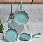 Juego de sartenes SANKEY 3 piezas color verde turquesa (16 cm, 20 cm y 26 cm) con recubrimiento antiadherente tipo marble coating; una sartén colgada al centro, otra apoyada inclinada a la izquierda y una sartén pequeña sobre la encimera a la derecha, con espátula negra colgada y planta decorativa.