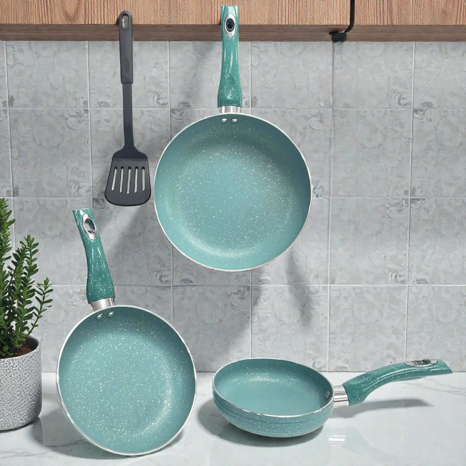 Juego de sartenes SANKEY 3 piezas color verde turquesa (16 cm, 20 cm y 26 cm) con recubrimiento antiadherente tipo marble coating; una sartén colgada al centro, otra apoyada inclinada a la izquierda y una sartén pequeña sobre la encimera a la derecha, con espátula negra colgada y planta decorativa.
