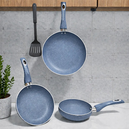 Juego de sartenes SANKEY color azul acero, set 3 piezas (16 cm, 20 cm y 26 cm) con recubrimiento antiadherente tipo marble coating; una sartén grande colgada al centro, otra apoyada inclinada a la izquierda y una sartén pequeña sobre la encimera a la derecha, con espátula negra colgada y planta decorativa.