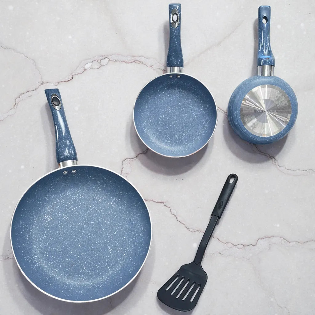Vista superior flat lay del set de sartenes SANKEY azul acero (3 piezas: 16/20/26 cm) antiadherentes tipo marble coating; se observan dos sartenes por dentro y una sartén volteada mostrando la base metálica circular, junto a una espátula negra sobre fondo tipo mármol.
