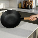 Wok Sankey de hierro fundido negro de 30 cm (12 pulgadas) con mango de madera, interior con superficie tipo panal, sostenido con la mano en cocina.