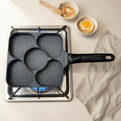 Sarten multifuncional Synamore 5 en 1 de color gris moteado con mango negro, colocado sobre estufa de gas en vista superior, con cinco compartimentos para cocinar huevo, pancakes y desayuno por separado.