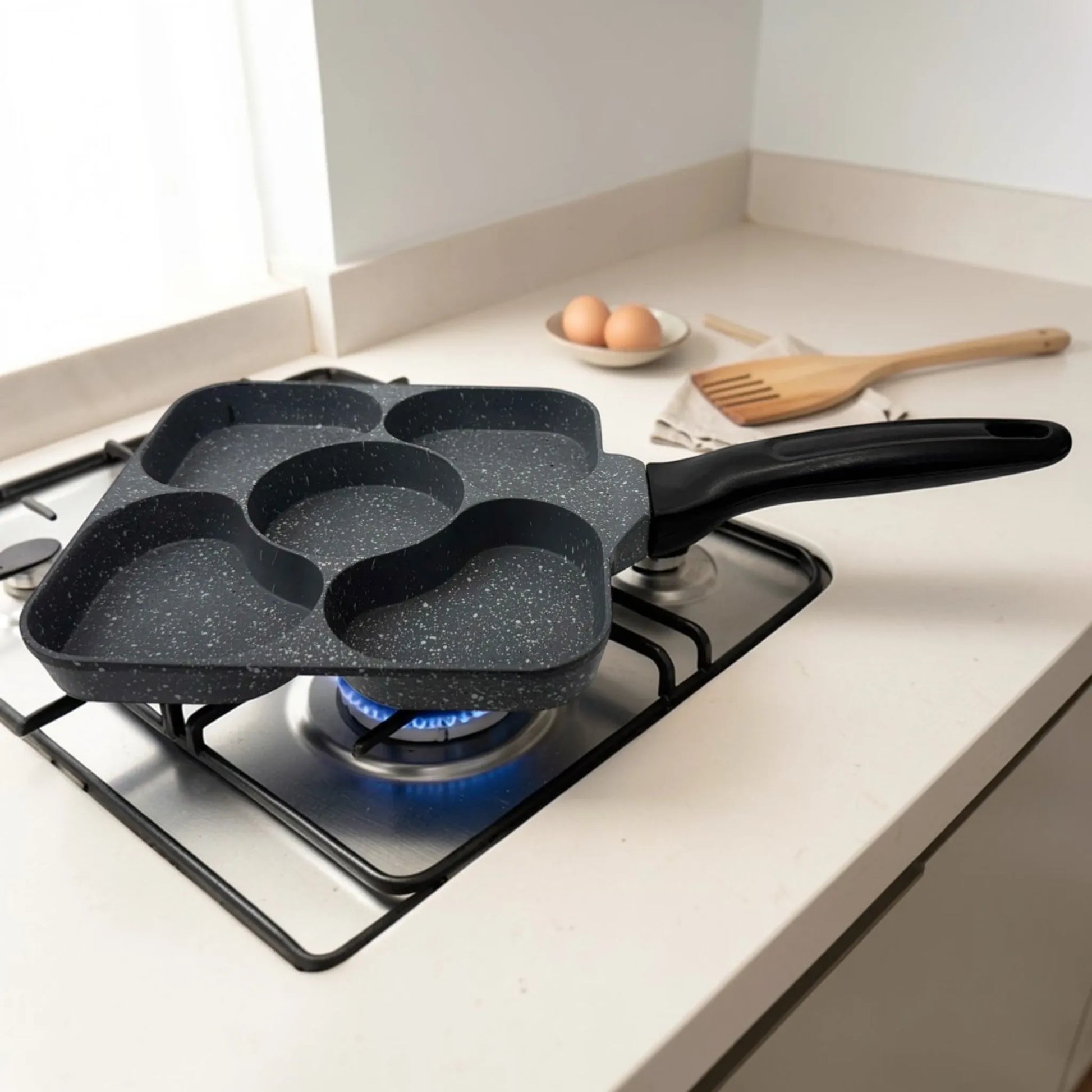 Sarten cuadrado Synamore 5 en 1 antiadherente en color gris moteado con mango negro, fotografiado en ángulo sobre una estufa de gas, mostrando sus cinco secciones de coccion separada.