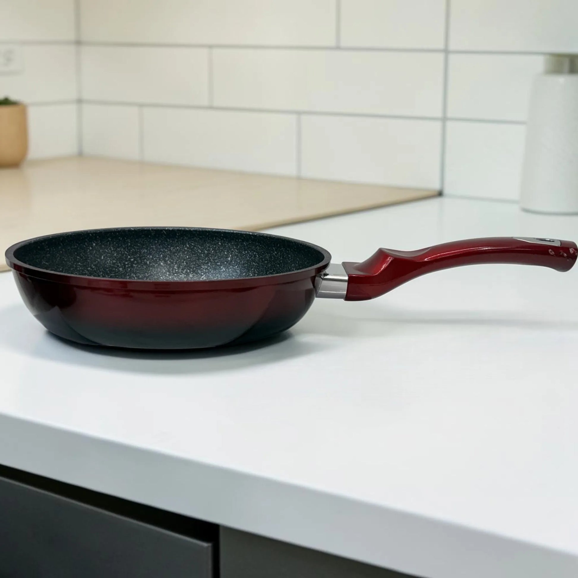 Sartén Epicure Escarlata en color rojo vino con interior antiadherente tipo mármol y mango largo ergonómico, fotografiada de lado sobre encimera blanca.