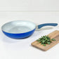 Sartén ROCA azul sin tapa con interior cerámico blanco, mango ergonómico y tabla de cocina decorativa al lado.