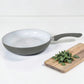 Sartén ROCA gris sin tapa con interior cerámico blanco, mango ergonómico y tabla de cocina decorativa al lado.