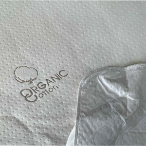 Detalle de la tela con textura y logo Organic Cotton del protector de colchón Scarlett Classics, mostrando la membrana impermeable en la esquina.