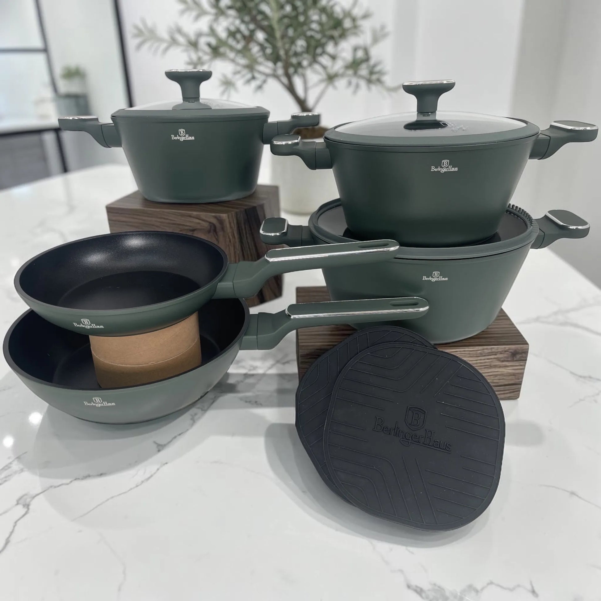 Set de cocina Berlinger Haus Matte Green con ollas, sartenes, tapas de vidrio templado y tapetes de silicona sobre encimera de mármol.