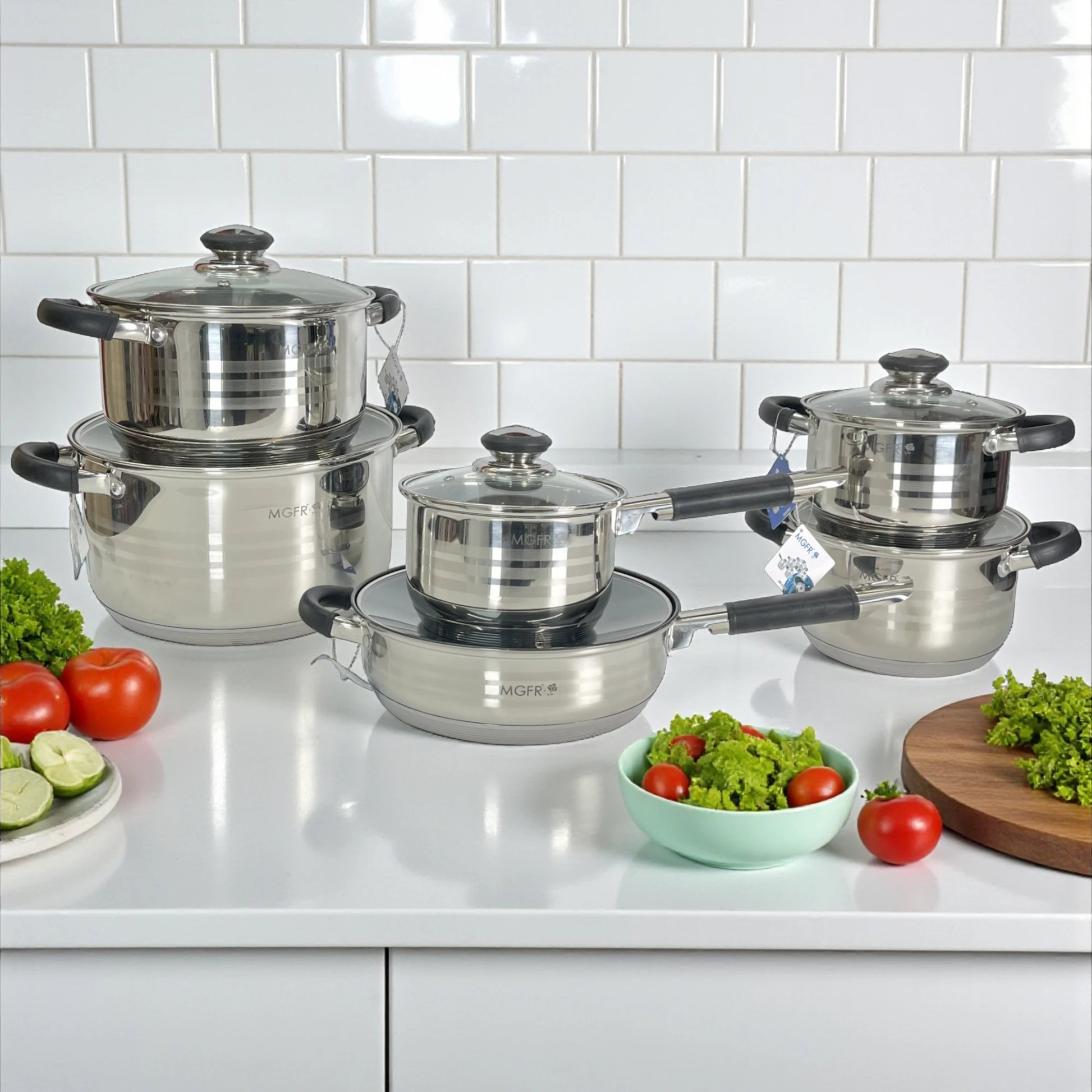 Set de ollas MGFR MR-290G 12 piezas en acero inoxidable 18/10; cacerolas 16, 18, 20 y 24 cm apiladas, sartén 24 cm y cazo 16 cm con tapas de vidrio en cocina blanca.
