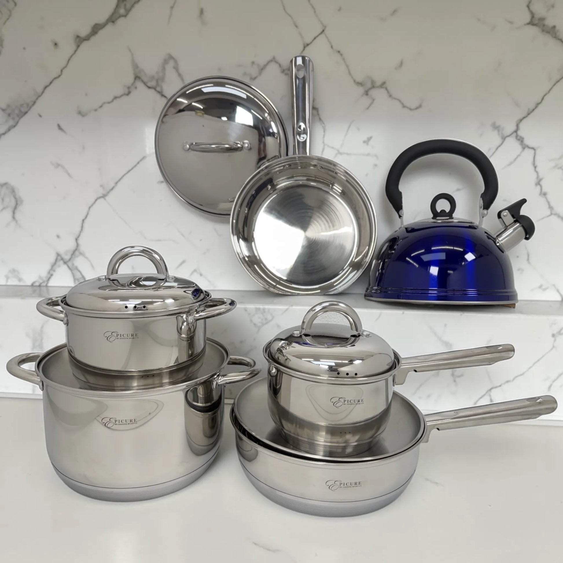 Set de ollas de acero inoxidable Epicure by Joseph Marcus con tetera azul y sartenes organizados sobre superficie de mármol en cocina moderna.