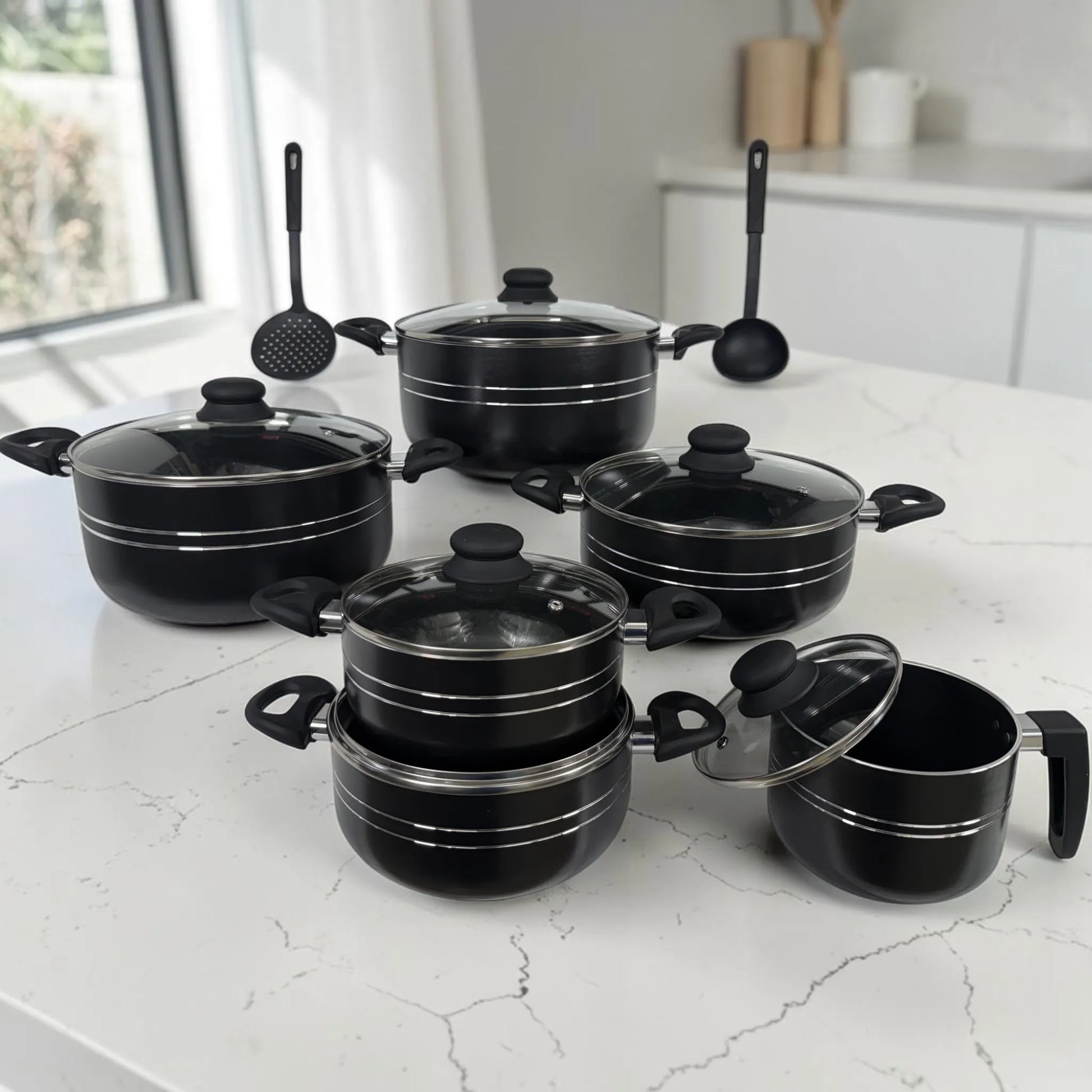 Set de ollas ROCA negro de 12 piezas antiadherente exhibido sobre cocina moderna con tapas de vidrio y utensilios.