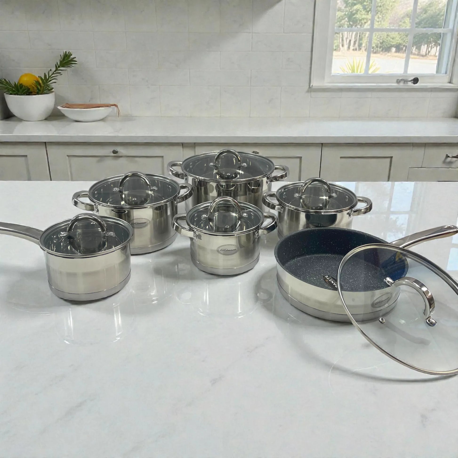 Set completo de ollas Rönner Austria de 12 piezas en acero inoxidable con tapa de vidrio y sartén granito, exhibido sobre mesón de cocina.