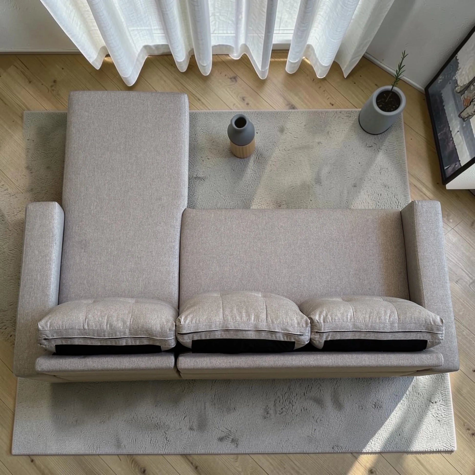 Vista superior del sofá cama 2 en 1 Ambienta Home Decor color gris claro con chaise longue, tres cojines removibles, brazos rectos y tapizado tipo lino en sala moderna.