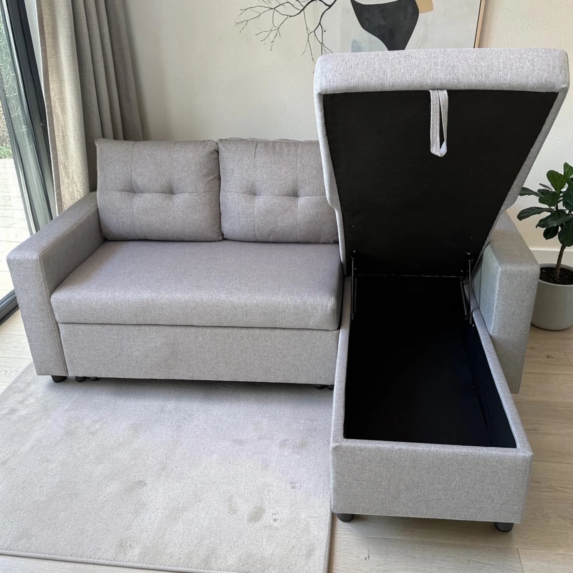 Sofá cama con cajón de almacenamiento color gris claro, vista frontal con compartimento abierto, tapa elevada e interior negro para guardar objetos.