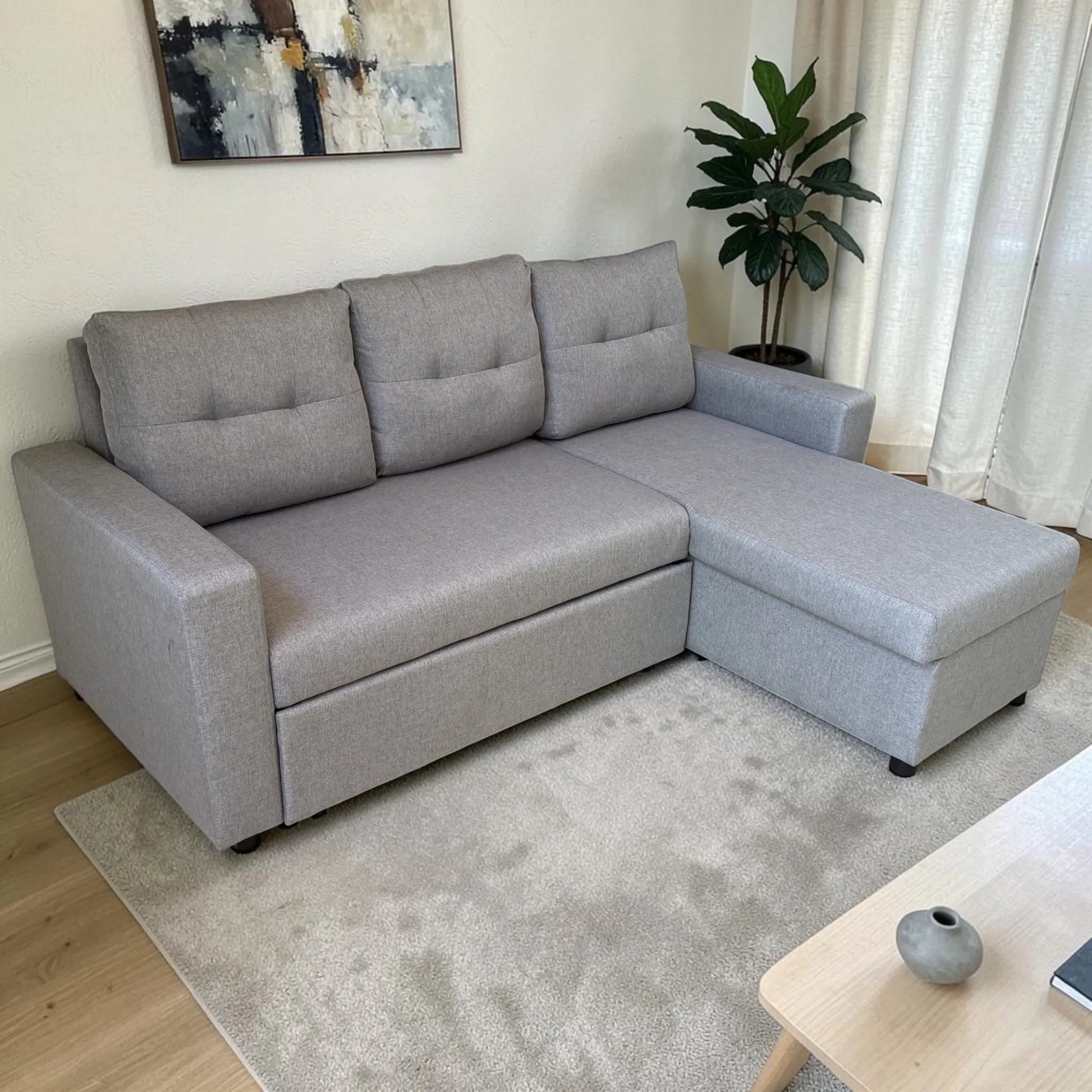 Sofá cama con cajón de almacenamiento Ambienta Home Decor color gris claro, vista lateral en sala moderna, diseño tipo chaise longue con brazos rectos y respaldos acolchados.