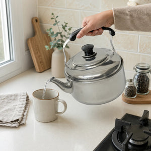 Persona sirviendo agua desde una tetera Lotus de acero inoxidable de 6 litros en una taza, mostrando el mango ergonómico y la boquilla vertedora.
